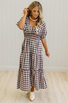 A woman wearing a brown and white gingham tiered maxi dress with puff sleeves and a smocked waist, standing in a room with light wood floors and white paneled walls.