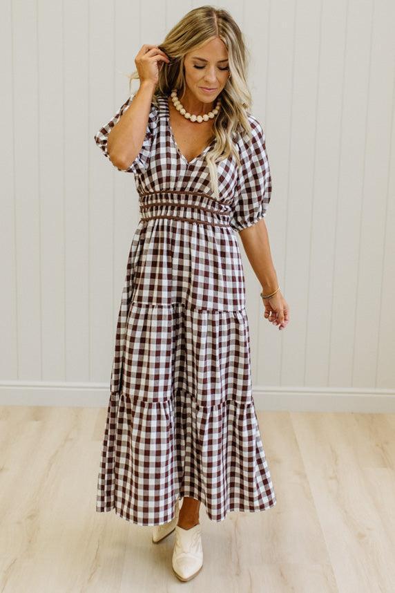 A woman wearing a brown and white gingham tiered maxi dress with puff sleeves and a smocked waist, standing in a room with light wood floors and white paneled walls.