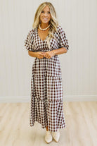 A woman modeling a brown and white gingham tiered maxi dress with puff sleeves, standing in front of a white paneled wall and light wood floor.