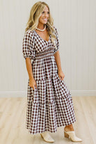 A woman modeling a brown and white gingham tiered maxi dress with puff sleeves, paired with cream-colored mule shoes and a pearl necklace, standing in front of a white paneled wall and light wood floor.