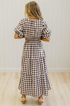 A woman wearing a brown and white gingham tiered maxi dress with puff sleeves and a smocked waist, standing in a room with light wood floors and white paneled walls.
