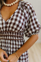 A close-up of a woman wearing a brown and white gingham dress with puff sleeves and a smocked waist, accessorized with a pearl necklace and gold bracelets.
