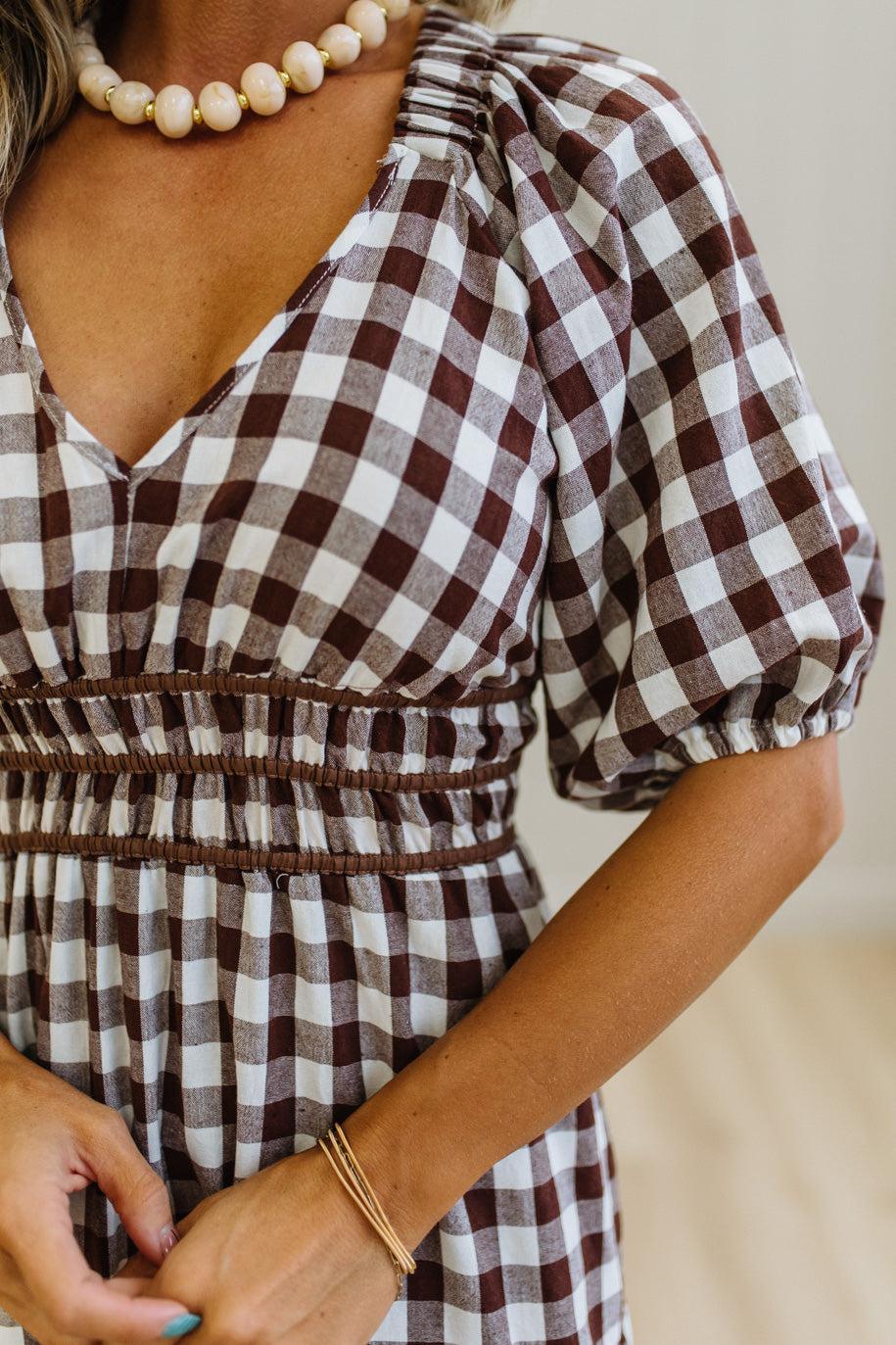 A close-up of a woman wearing a brown and white gingham dress with puff sleeves and a smocked waist, accessorized with a pearl necklace and gold bracelets.