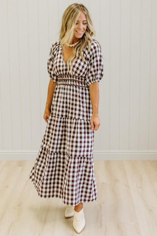 A woman wearing a brown and white gingham tiered maxi dress with puff sleeves and a smocked waist, standing in a room with light wood floors and white paneled walls.