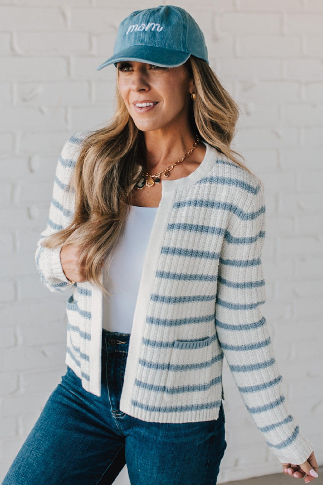 Blonde woman in a denim hat wear a cream and blue striped sweater cardigan with pockets