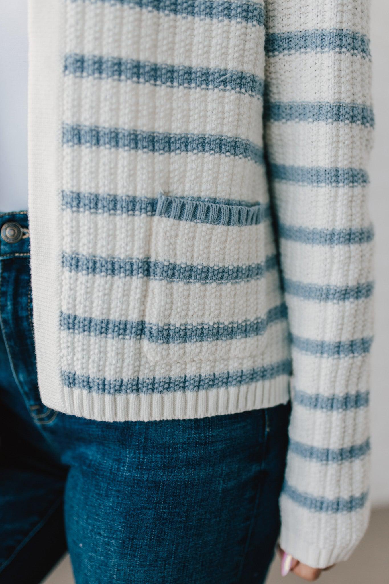 Blonde woman in a denim hat wear a cream and blue striped sweater cardigan with pockets