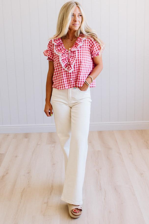 Woman wearing a red and white checkered blouse with ruffles and white pants in a room with wooden flooring.