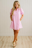 Woman wearing a pink checkered dress standing against a white paneled wall.