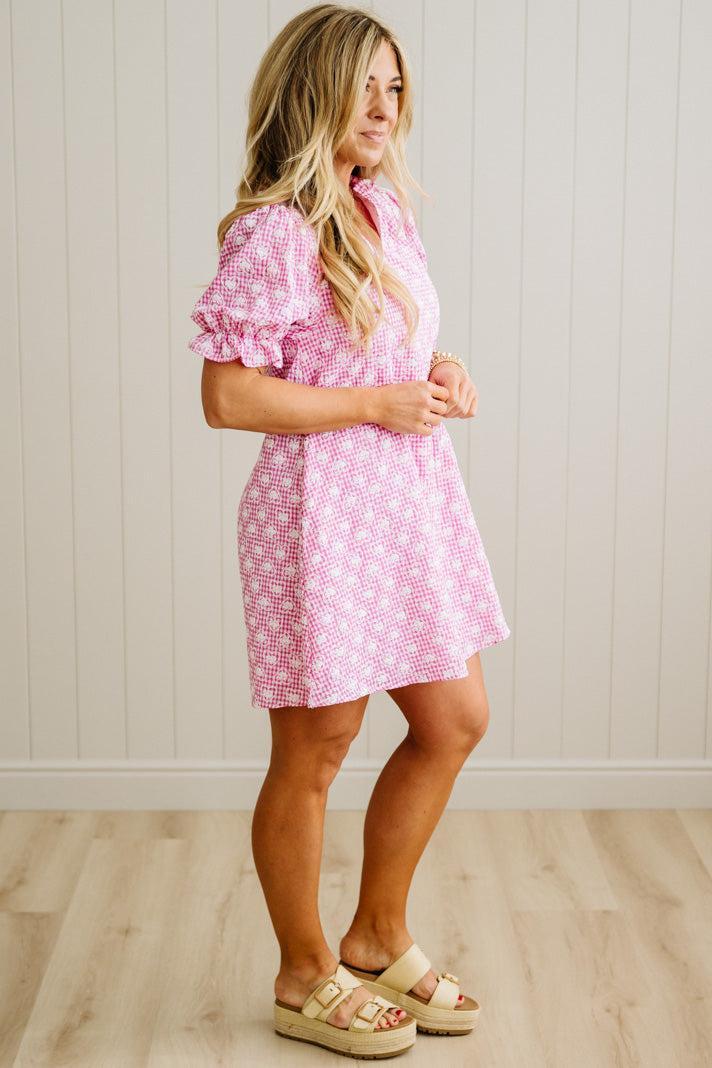 Woman wearing a pink checkered dress standing against a white paneled wall.