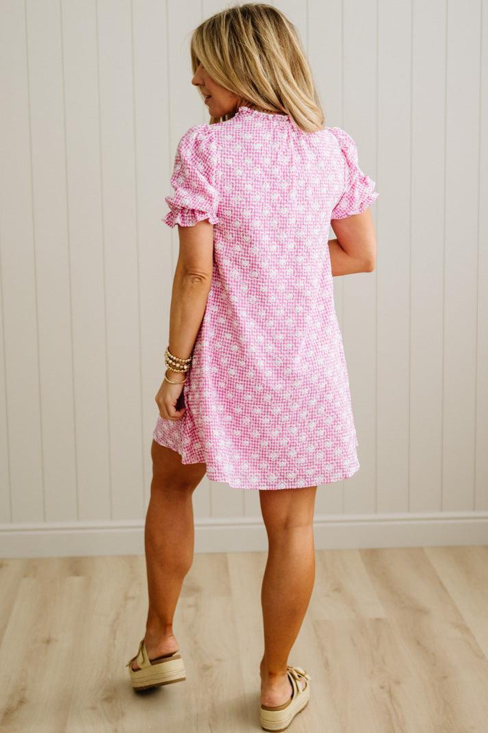 Woman wearing a pink checkered dress standing against a white paneled wall.