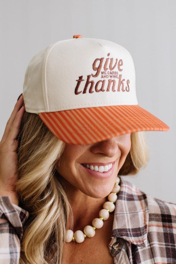 Blonde smiling woman wearing a cream cap with striped orange brim and embroidered text on the front that says "Give me carbs and wine, thanks" at the center.