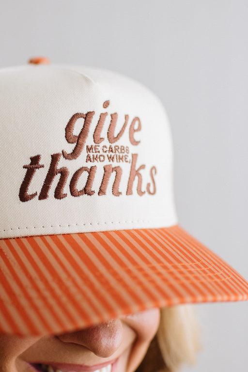 Blonde smiling woman wearing a cream cap with striped orange brim and embroidered text on the front that says "Give me carbs and wine, thanks" at the center.