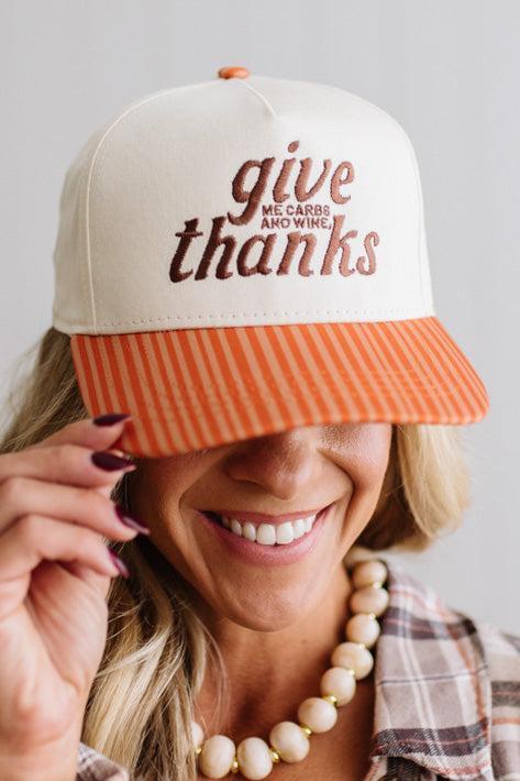 Blonde smiling woman wearing a cream cap with striped orange brim and embroidered text on the front that says "Give me carbs and wine, thanks" at the center.