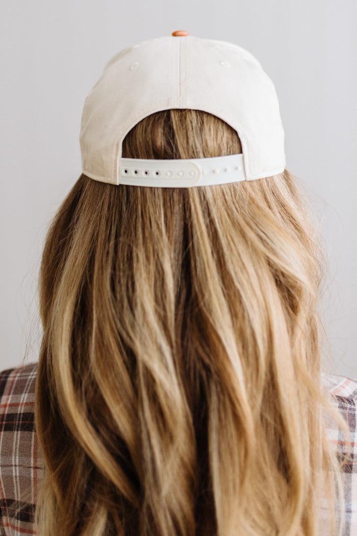 Blonde smiling woman wearing a cream cap with striped orange brim and embroidered text on the front that says "Give me carbs and wine, thanks" at the center.