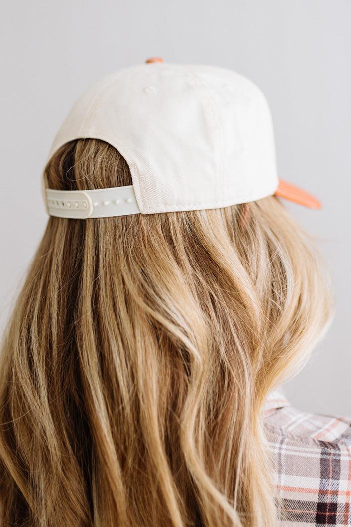Blonde smiling woman wearing a cream cap with striped orange brim and embroidered text on the front that says "Give me carbs and wine, thanks" at the center.