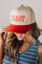 Blonde woman wearing a two tone cream and red trucker hat with GOD BLESS AMERICA embroidered at the front.