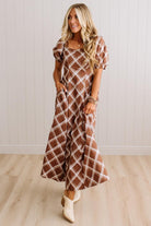 A woman modeling a brown and white plaid maxi dress with puff sleeves and side pockets, standing in a room with light wood floors and white paneled walls.