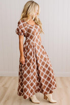 A woman modeling a brown and white diamond-patterned maxi dress with puff sleeves, paired with beige ankle boots, standing in front of a white paneled wall and light wood floor.