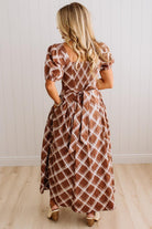 A woman viewed from behind wearing a brown and white diamond-patterned midi dress with puff sleeves and a tie waist, standing on a light wood floor against a white paneled wall.