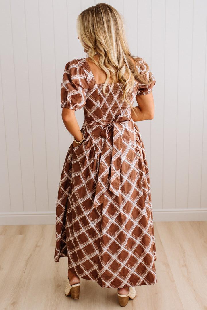 A woman viewed from behind wearing a brown and white diamond-patterned midi dress with puff sleeves and a tie waist, standing on a light wood floor against a white paneled wall.
