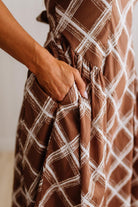 Close-up of a brown dress with a white plaid pattern, featuring a side pocket into which a hand is placed.