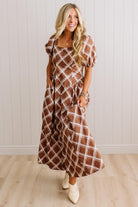 A woman modeling a brown and white plaid midi dress with puff sleeves and side pockets, standing in front of a white paneled wall and light wood floor.