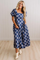 A woman modeling a navy blue midi dress with a white plaid pattern, featuring puff sleeves, a square neckline, and side pockets, paired with white cowboy boots against a white paneled wall and light wood floor.