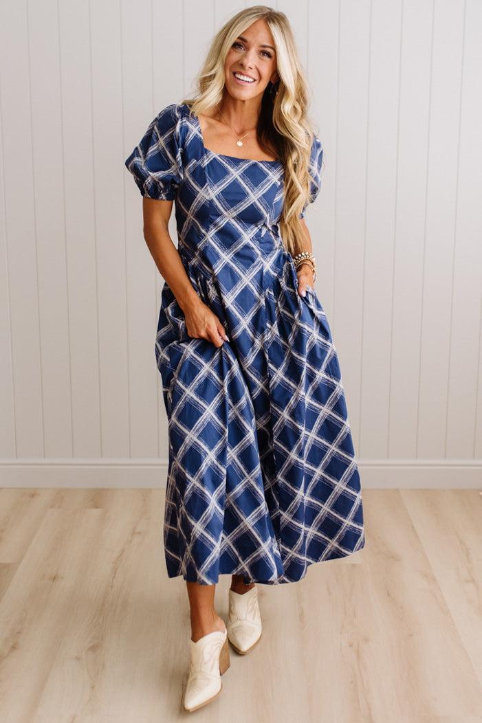 A woman modeling a navy blue midi dress with a white plaid pattern, featuring puff sleeves, a square neckline, and side pockets, paired with white cowboy boots against a white paneled wall and light wood floor.