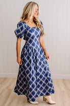 A woman wearing a blue and white plaid maxi dress with puff sleeves, standing in a room with light wood floors and white paneled walls.
