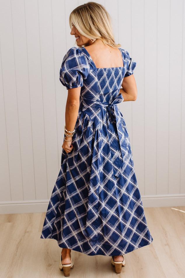 A woman wearing a navy blue maxi dress with a white plaid pattern, shown from the back. The dress features puff sleeves, a square neckline, and a tie at the waist.