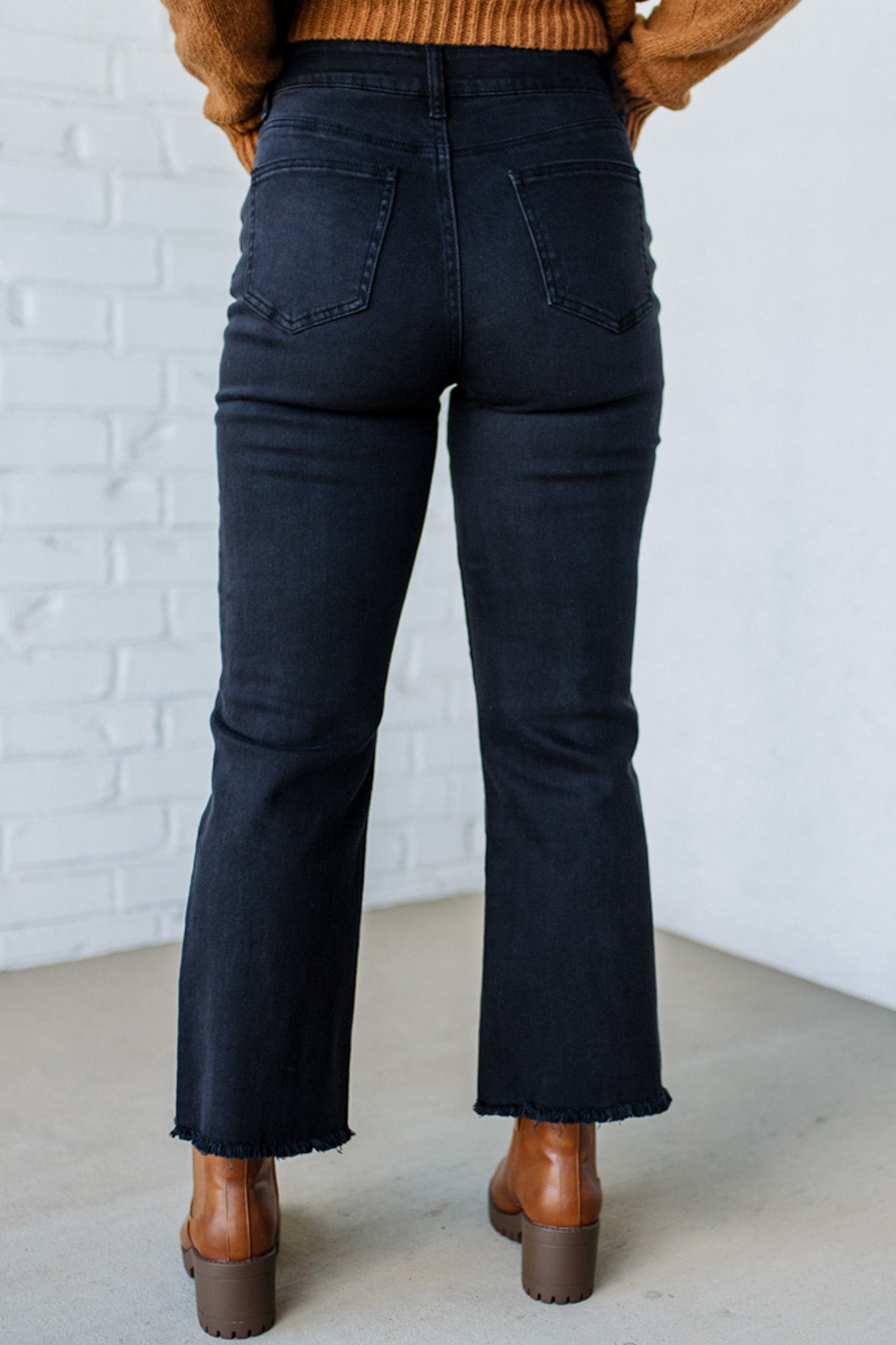 Person wearing black flared jeans and brown boots against a white brick wall.