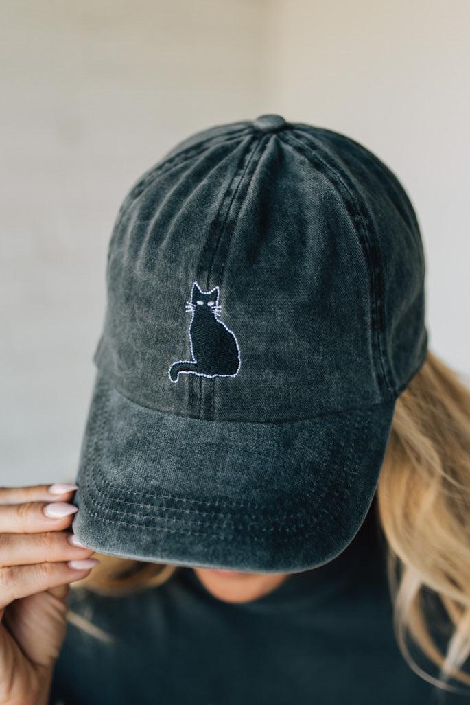 Black cap with a cat design held by a person against a neutral background