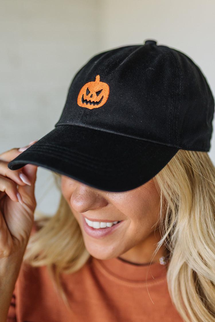 Black cap with orange pumpkin design worn by a person, against a neutral background