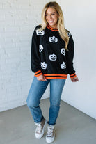 Woman wearing a black Halloween-themed sweater with white pumpkins and orange trim, standing against a white brick wall.