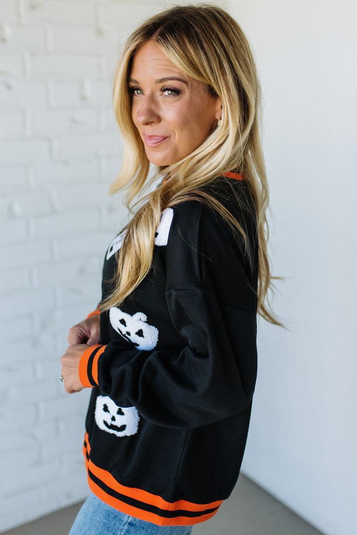 Woman wearing a black Halloween-themed sweater with white pumpkins and orange accents against a white brick wall.