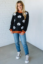 Woman wearing a black sweater with white pumpkin patterns, blue jeans, and white sneakers against a white brick wall.