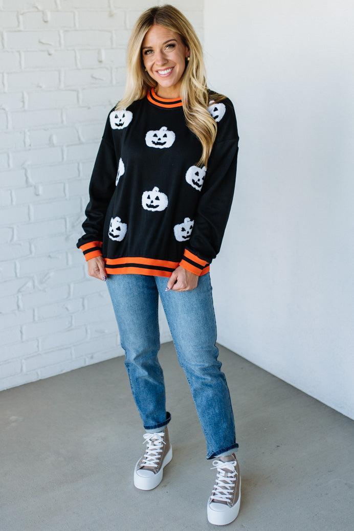 Woman wearing a black sweater with white pumpkin patterns, blue jeans, and white sneakers against a white brick wall.