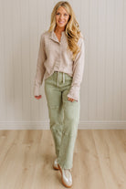 Woman wearing a light-colored shirt and green pants standing against a plain wall.