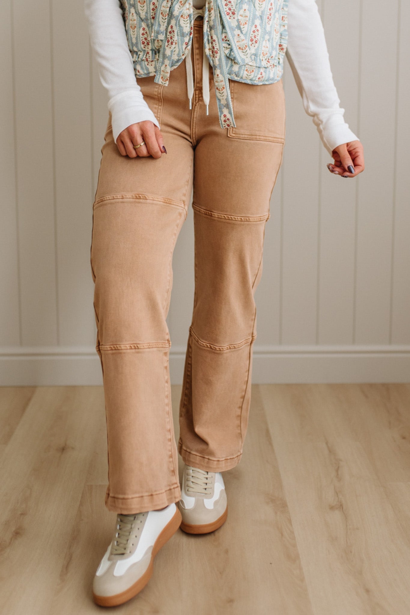 Person wearing beige pants and a floral top in a room with wooden flooring and a neutral wall.