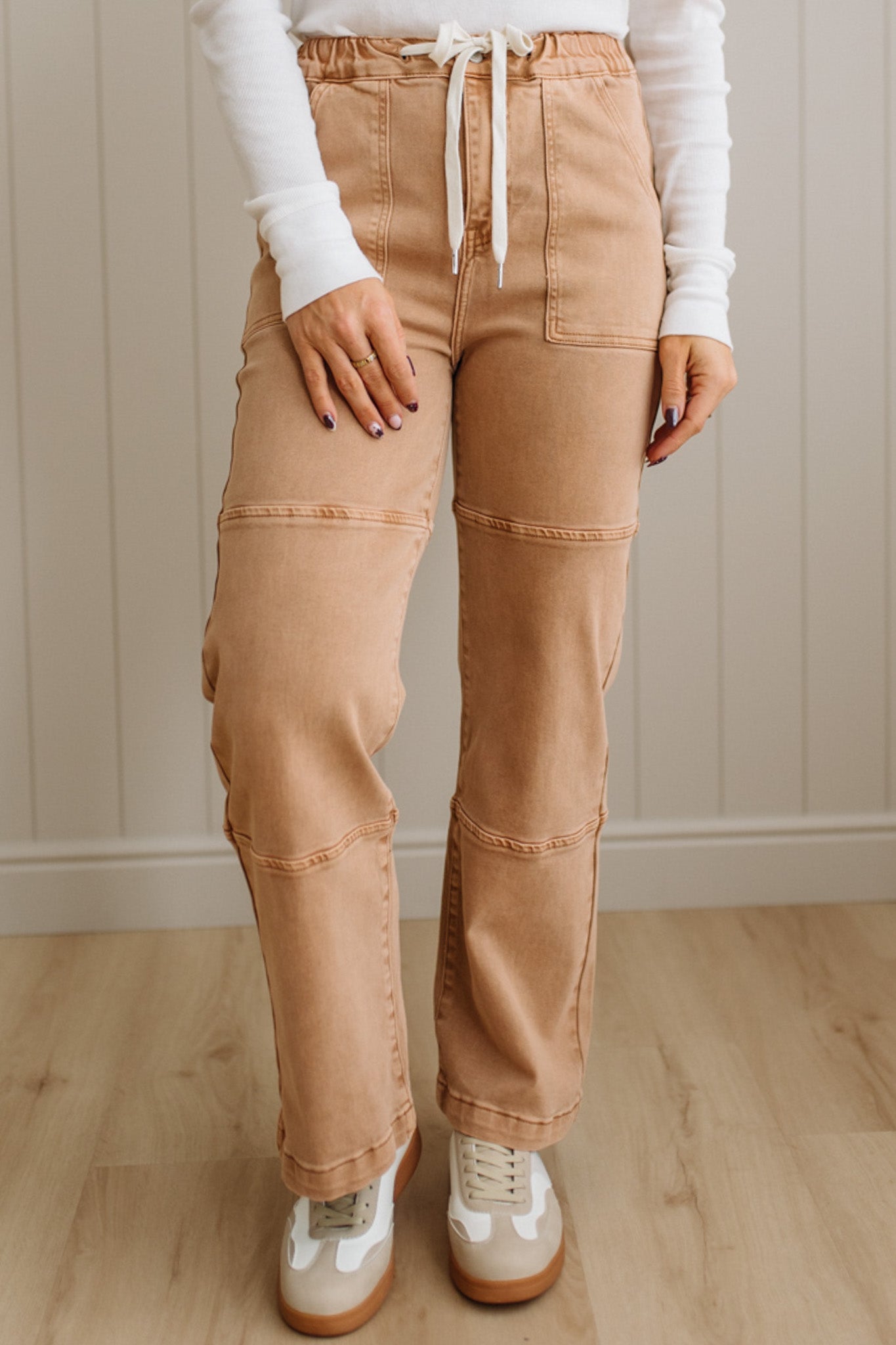Person wearing tan pants with a white shirt and white shoes in a room with wooden flooring and a neutral wall.