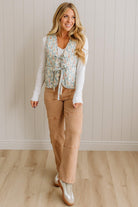 Woman wearing a floral vest, white long-sleeve shirt, tan pants, and silver shoes against a beige paneled wall.
