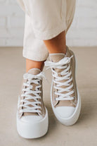 Beige high-top sneakers with white laces and soles worn with light-colored pants on a neutral background.