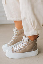 Beige high-top sneakers with white laces worn with light-colored pants on a neutral background