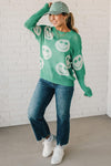 BLONDE WOMEN IN A GREEN AND WHITE CHECKER HAT WEARING A GREEN AND WHITE LOW GAUGE SWEATER WITH SMILEY WITH SHAMROCK EYES PATTERN.