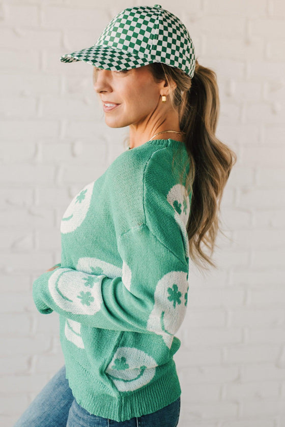 BLONDE WOMEN IN A GREEN AND WHITE CHECKER HAT WEARING A GREEN AND WHITE LOW GAUGE SWEATER WITH SMILEY WITH SHAMROCK EYES PATTERN.