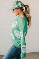 BLONDE WOMEN IN A GREEN AND WHITE CHECKER HAT WEARING A GREEN AND WHITE LOW GAUGE SWEATER WITH SMILEY WITH SHAMROCK EYES PATTERN.