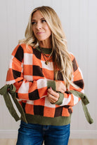 Blonde woman wearing a orange, brown and cream check sweater with olive green ribbed trim and bows at the elbow.