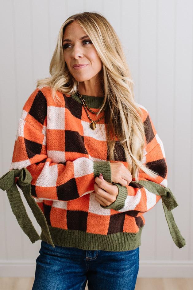 Blonde woman wearing a orange, brown and cream check sweater with olive green ribbed trim and bows at the elbow.