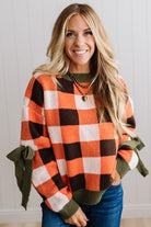 Blonde woman wearing a orange, brown and cream check sweater with olive green ribbed trim and bows at the elbow.