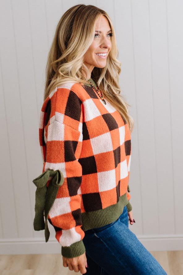 Blonde woman wearing a orange, brown and cream check sweater with olive green ribbed trim and bows at the elbow.